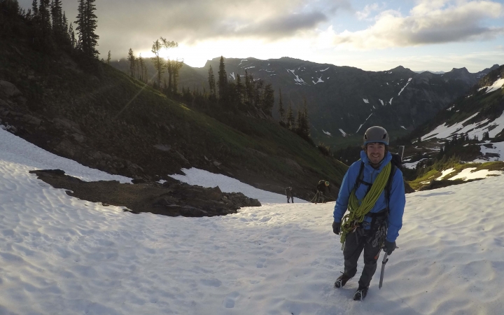 A person wearing safety gear stands on the snow in front of a vast mountainous landscape